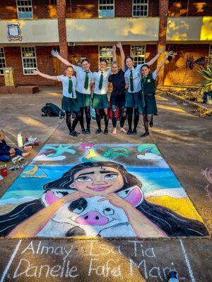 Six students in school uniforms stand on a sunny pavement, smiling with arms raised. Below them is a vibrant chalk art of a girl and pig. The atmosphere is joyful.