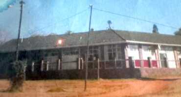A single-story brick building with large windows and a gabled roof is seen under a clear blue sky. In front, a dirt path and a grassy area are visible.