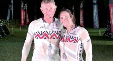 A man and woman smiling, wearing matching cycling jerseys with geometric patterns and medals. They stand on grass at night, with banners in the background.