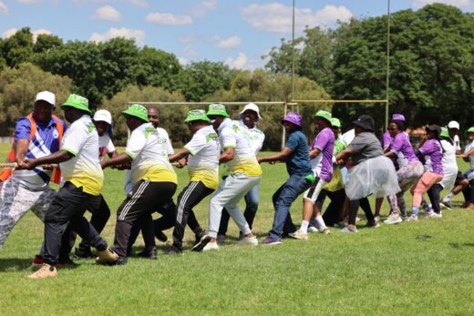 A diverse group of people playing tug-of-war on a grassy field, dressed in colorful casual attire and hats, under a bright blue sky with scattered clouds.