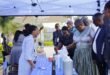 People gather under a canopy at an outdoor market stall displaying cleaning products. The scene is lively, with attendees engaged and interacting.