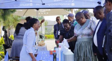 People gather under a canopy at an outdoor market stall displaying cleaning products. The scene is lively, with attendees engaged and interacting.