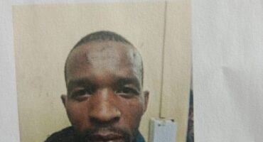 A person wearing a dark hoodie looks directly at the camera with a neutral expression. The background is an off-white wall with a device attached.