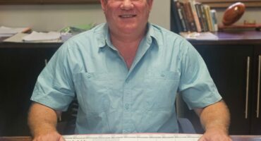 A man in a light blue shirt sits at a desk with a September 2023 calendar. Behind him are brown blinds, shelves with books, and a decorative item. The setting is calm.