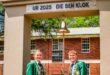 Two smiling students in green blazers stand under an arch labeled "UR 2025 DIE BEN KLOK," with a brass bell. A red-brick building is behind them.