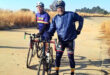 Two cyclists in helmets and cycling gear pause on a sunlit path surrounded by tall, dry grass. They smile, conveying a sense of camaraderie.
