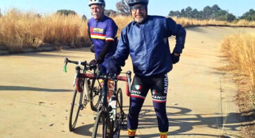 Two cyclists in helmets and cycling gear pause on a sunlit path surrounded by tall, dry grass. They smile, conveying a sense of camaraderie.