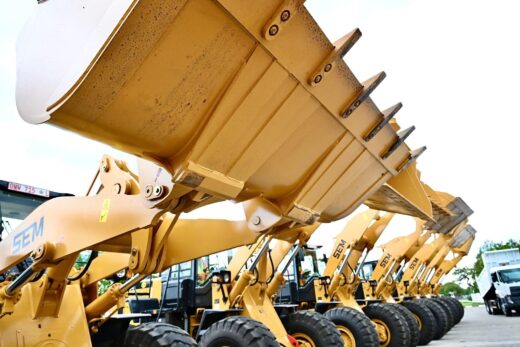 A row of yellow construction loaders with raised buckets lines up on a paved ground. Overcast sky and greenery in the background suggest an industrial setting.