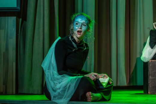 Young woman in blue face paint and black outfit sits on stage, looking surprised. She wears a translucent shawl. Green lighting and curtains in background.