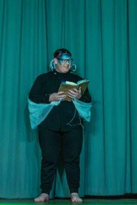 Person on stage, dressed in black with blue headpiece and sheer fabric wings, reads a book while smiling. Green curtain backdrop, theatrical setting.