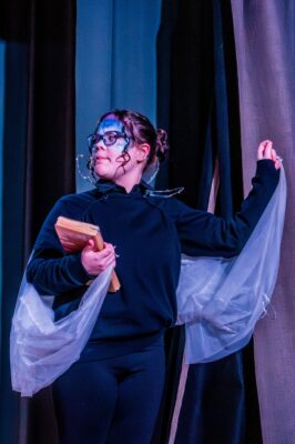 A person on stage wears a dark outfit with sheer, wing-like fabric, glasses, and blue face paint. They hold a book, exuding a theatrical, mysterious vibe.
