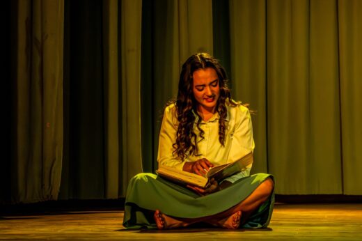 A woman sits cross-legged on a stage, reading an open book. She's wearing a long skirt and blouse. The scene is warmly lit, creating a serene atmosphere.