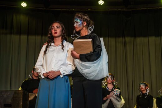 Two women on stage during a theater performance; one in a white blouse looks solemn, while the other, with face paint and a costume, offers comfort.