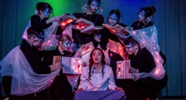 A group of performers with painted faces and glowing books encircle a seated woman in white and blue, creating a mystical, theatrical scene.