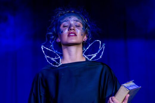 A performer in a theatrical setting, with glittering face paint and futuristic blue lighting, holds a book and closes eyes in a serene, dramatic pose.
