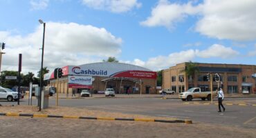 Street view of a Cashbuild store under a partly cloudy sky. Cars and a person are crossing the intersection. The traffic light is not in working order. The mood is bright and bustling.