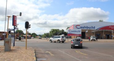 A sunny day at an intersection with cars driving near a Cashbuild store. Traffic lights are visible, but not working. The sky is partly cloudy, creating a calm atmosphere.