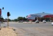 Street intersection with a traffic light that is out of order, clear blue sky, and a large Cashbuild hardware store on the right. Vehicles are parked outside the store.