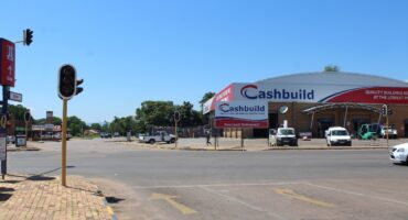 Street intersection with a traffic light that is out of order, clear blue sky, and a large Cashbuild hardware store on the right. Vehicles are parked outside the store.