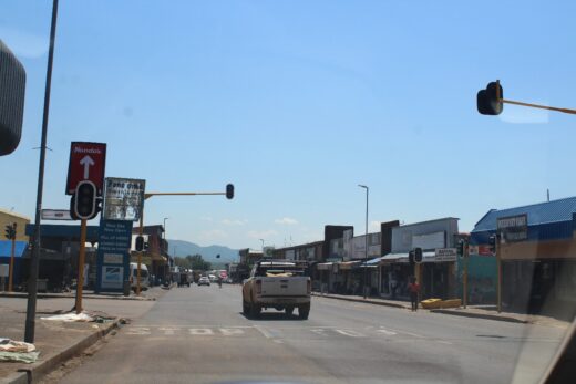 Street view with traffic lights that’s not in working order, a white pickup truck in the center, and shops lining both sides. A Nando's sign is visible. Clear sky above.
