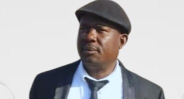 A man in a black beret and suit gazes thoughtfully to the side against a plain background, conveying a calm and introspective mood.