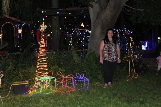 A woman stands beside colorful Christmas decorations under a tree at night. The festive lights include a tree and presents, creating a joyful atmosphere.