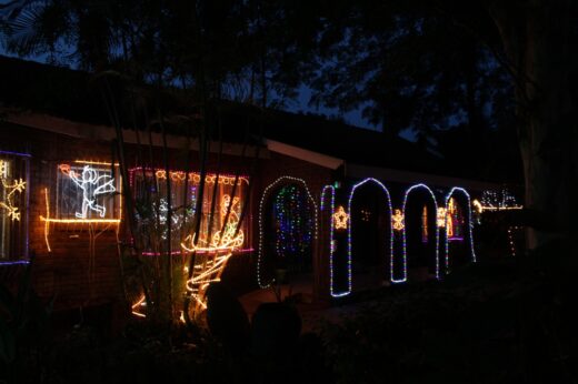 Nighttime scene of a house with vibrant holiday lights. Neon figures and arches glow in blue, orange, and white, creating a festive atmosphere.
