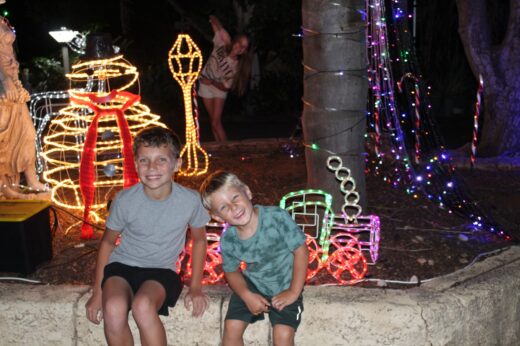 Two smiling boys sit on a stone ledge surrounded by colorful holiday lights shaped like gifts and a train. A playful person poses in the background.