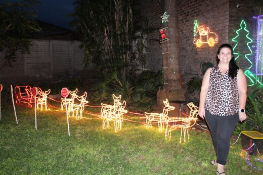 A woman stands smiling beside glowing Christmas decorations, including a sleigh and reindeer made of lights on a lawn. The scene feels festive and cheerful.