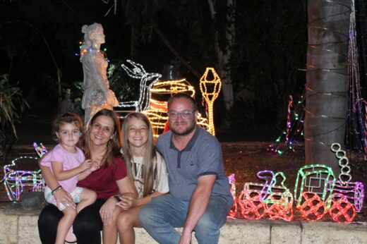 A family of four, two adults and two children, sit on a low wall at night, smiling. Behind them are colorful holiday lights and sculptures, creating a festive atmosphere.