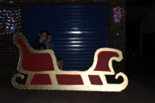 A girld and boy sit on a festive red and gold sleigh adorned with colorful lights. A blue garage door and holiday decor are in the background.
