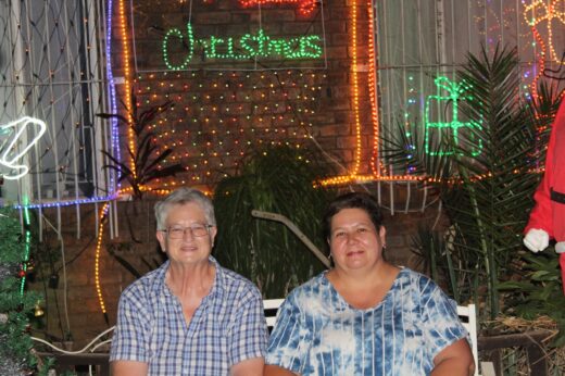 Two people smiling in front of colorful Christmas lights and decorations, including a lit sign reading "Christmas" and a gift box. Festive, joyful atmosphere.