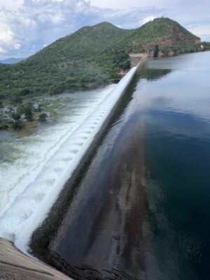 A large dam with cascading water on the left contrasts the calm reservoir on the right, set against lush green hills under a cloudy sky.