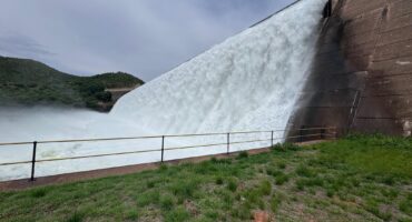 A dam releases a powerful torrent of water over its edge, cascading down in a white rush. Lush green grass and a safety railing are in the foreground.
