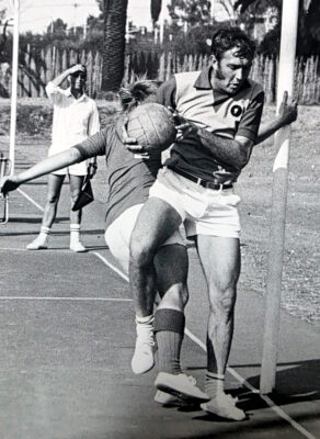 Black and white photo of two athletes in mid-action during a korfball game, with one holding the ball near a post. Background shows another person observing.