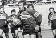 Black and white photo of rugby match many years ago. A determined player, holding the ball, runs ahead while teammates follow closely, exuding teamwork and intensity.