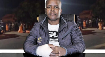 A man sits at a desk with a bandaged hand, wearing a dark jacket over a printed shirt. The background shows a nighttime scene with people gathered on a street, evoking a tense atmosphere.
