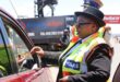 A police officer in a bright yellow vest hands back a document to a driver in a red car. The scene is outdoors with a busy road in the background.