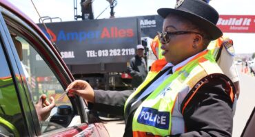 A police officer in a bright yellow vest hands back a document to a driver in a red car. The scene is outdoors with a busy road in the background.
