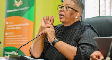 A person in glasses speaks passionately at a meeting, gesturing with their hands. Behind them is a banner with text and a colorful logo. The atmosphere is formal.
