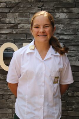 Smiling girl in a white school uniform stands against a stone wall. The uniform has a badge and logo. Her hair is tied back, conveying a friendly and confident tone.