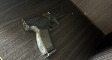 A black handgun lying on a dark wood-textured surface inside an open cabinet. The setting is dimly lit, conveying a serious and somber tone.