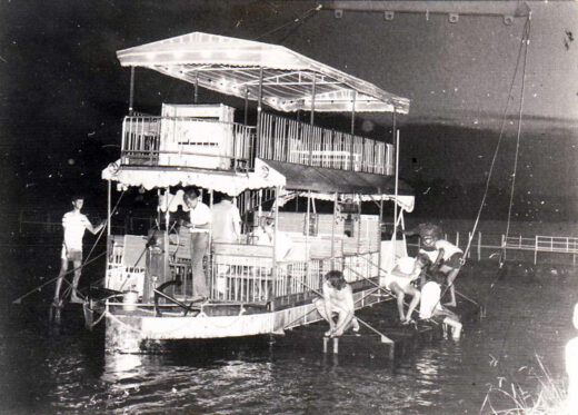 A vintage photo showing a two-story boat with several people on deck, navigating a body of water at night. Some are using poles for movement. The atmosphere is lively yet calm.