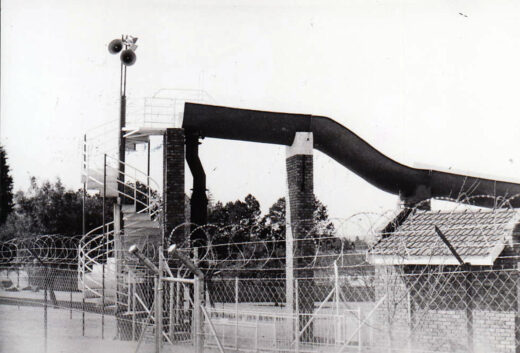 Black and white photo of an abandoned water slide beside a barbed wire fence, conveying a sense of desolation. A speaker tower and trees are in the background.