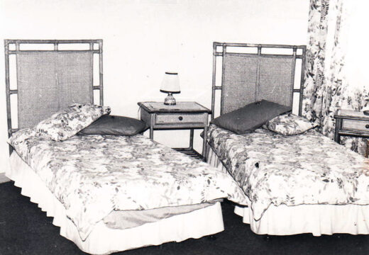 Two vintage twin beds with floral bedspreads are separated by a small wooden nightstand with a lamp. Floral curtains add a cozy, retro vibe.