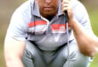 A golfer crouches on a green, focused with a putter in hand. He wears a gray shirt, cap, and watch, set against a blurred background, conveying concentration.