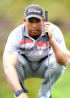 A golfer crouches on a green, focused with a putter in hand. He wears a gray shirt, cap, and watch, set against a blurred background, conveying concentration.