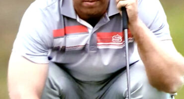 A golfer crouches on a green, focused with a putter in hand. He wears a gray shirt, cap, and watch, set against a blurred background, conveying concentration.