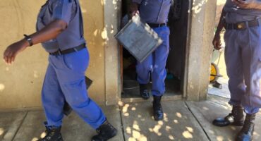 Three police officers in uniform outside a building entrance; one carries a case. The sunlight casts dappled shadows. The mood is serious.