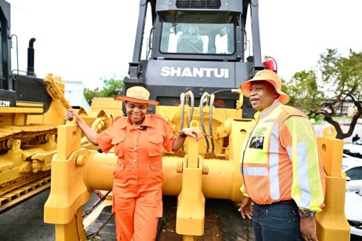 Two people in orange work attire stand in front of a large yellow Shantui bulldozer. They appear cheerful, with trees and cars in the background.
